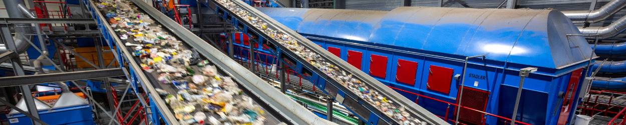 Afvalverwerkingsinstallatie met lopende banden waarop gemengd afval wordt getransporteerd, met op de achtergrond een blauwe industriële installatie of machinehal.