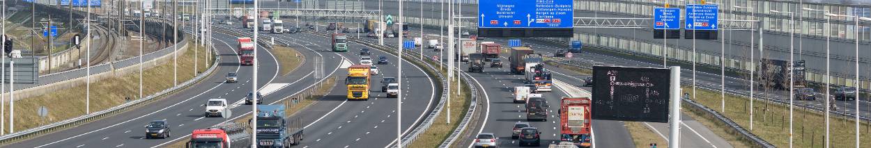 Drukke snelweg met meerdere rijstroken in beide richtingen, waarop vrachtwagens en personenauto’s rijden. Boven de weg hangen blauwe verkeersborden en een matrixbord met informatie voor weggebruikers. Links naast de weg ligt een spoorlijn