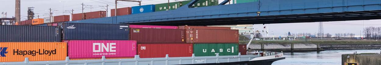 Vrachtschip met gestapelde zeecontainers vaart onder een blauwe brug door in een industriële havenomgeving. Op de achtergrond zijn spoorlijnen, kranen en elektriciteitsmasten zichtbaar.