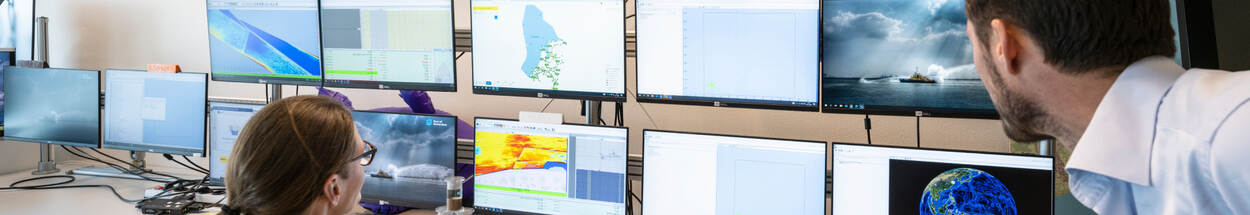 Twee mensen analyseren data op meerdere computerschermen met weerkaarten, satellietbeelden en geografische informatie in een monitoringruimte.