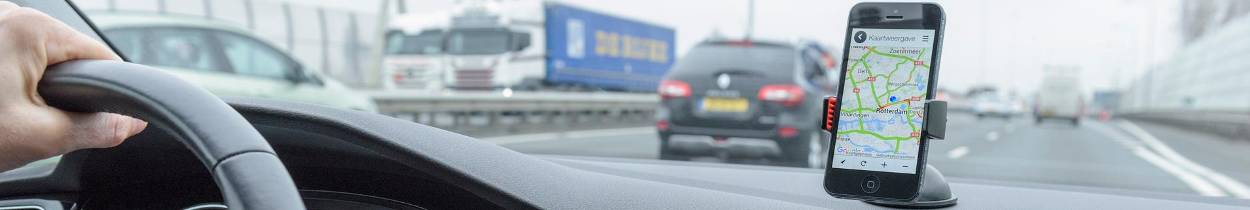 Bestuurder rijdt op een snelweg en gebruikt een navigatie-app op een smartphone die bevestigd is aan het dashboard. Andere auto's en vrachtwagens rijden op de weg, met geluidsschermen langs beide zijden.