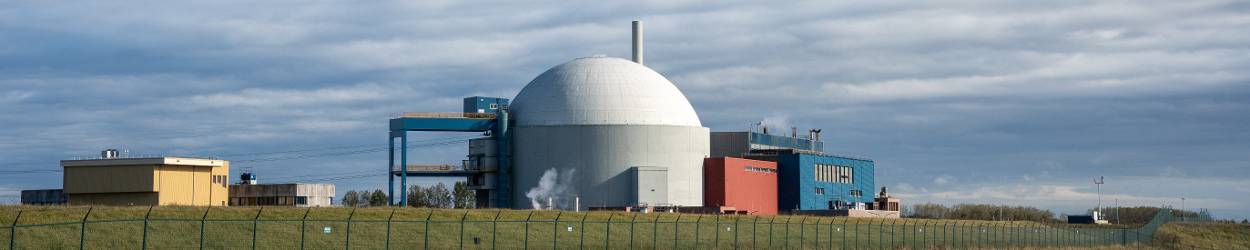 Kerncentrale met kenmerkende witte koepel en bijgebouwen, gelegen in een open landschap met hekwerk op de voorgrond en een bewolkte lucht op de achtergrond.