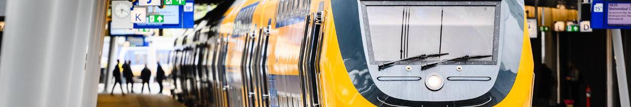 Een gele intercitytrein staat stil op een perron, met reizigers en informatieschermen zichtbaar op de achtergrond van het station
