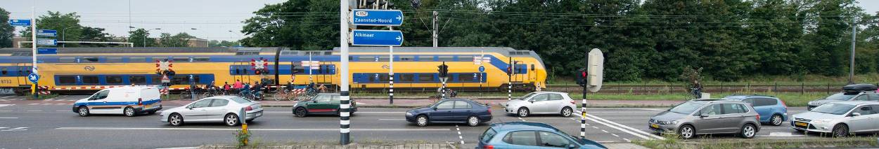 Drukke spoorwegovergang met wachtende auto's en fietsers terwijl een gele dubbeldekstrein van de NS passeert. Op de achtergrond groenstrook en bomen, op de voorgrond verkeersborden en kruispunt.