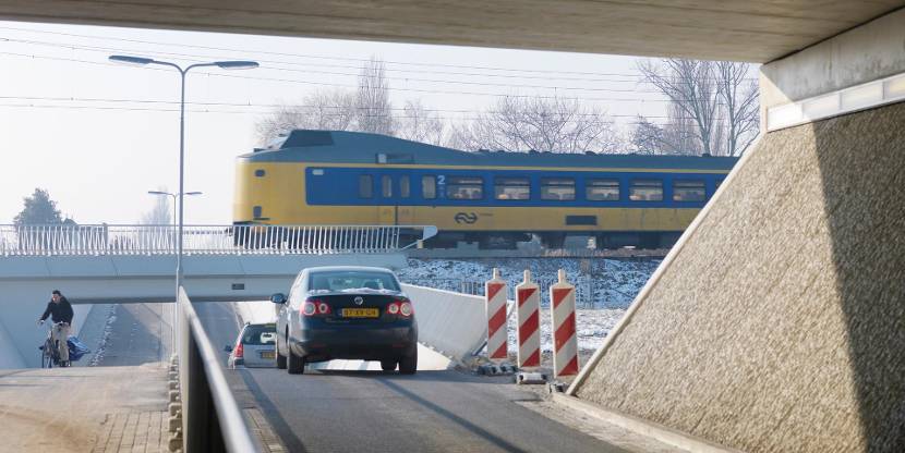 Trein van de NS rijdt over een spoorbrug terwijl auto's en een fietser onder het viaduct doorrijden; winterse omgeving met kale bomen op de achtergrond.