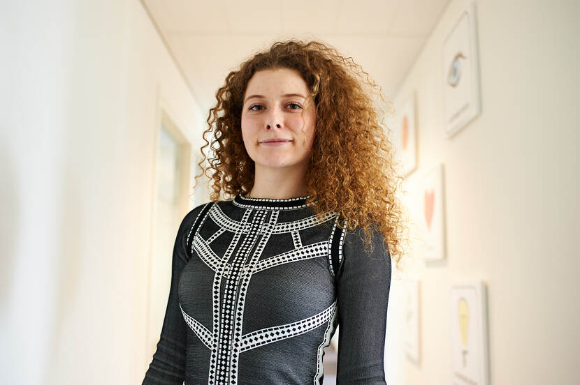 Robin Staa heeft lang, krullend roodbruin haar. Ze kijkt in de camera. Ze draagt een zwart shirt met een wit geometrisch patroon. De setting is een lichte gang met kunstwerken aan de muur.
