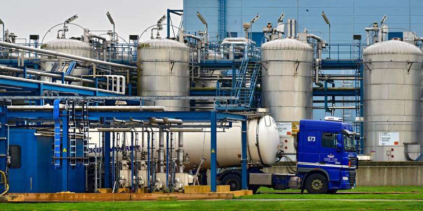 Industriële faciliteit met grote metalen opslagtanks en pijpleidingen, met een blauwe vrachtwagen met tankoplegger op de voorgrond.